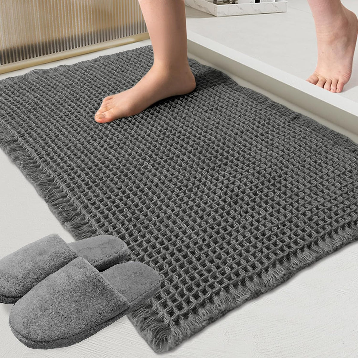 Close-up of a dark gray waffle bath mat with fringe showing texture and a bare foot stepping onto it; slippers nearby.