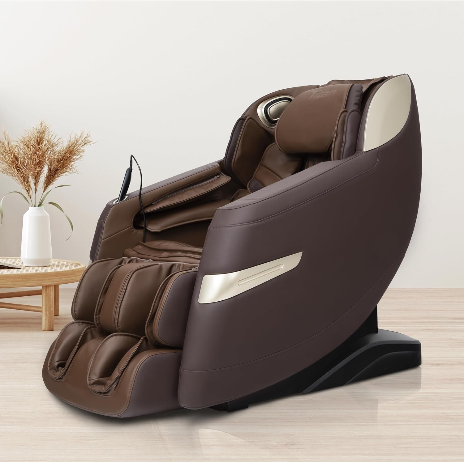 Three-quarter side view of a brown massage chair beside a low round table and vase on light wood flooring.
