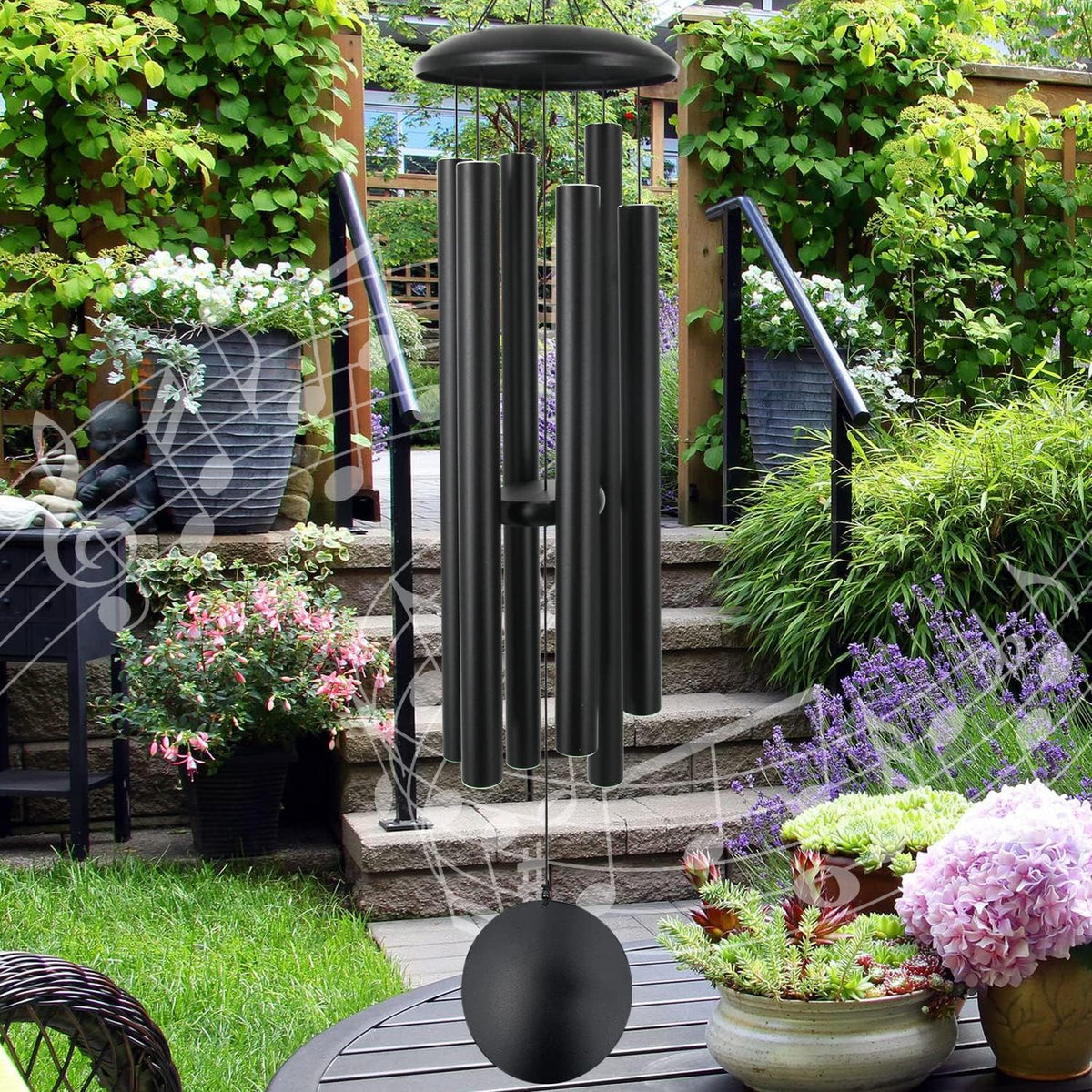 Black aluminum wind chime with six tubes and round sail hanging in a leafy garden, front view