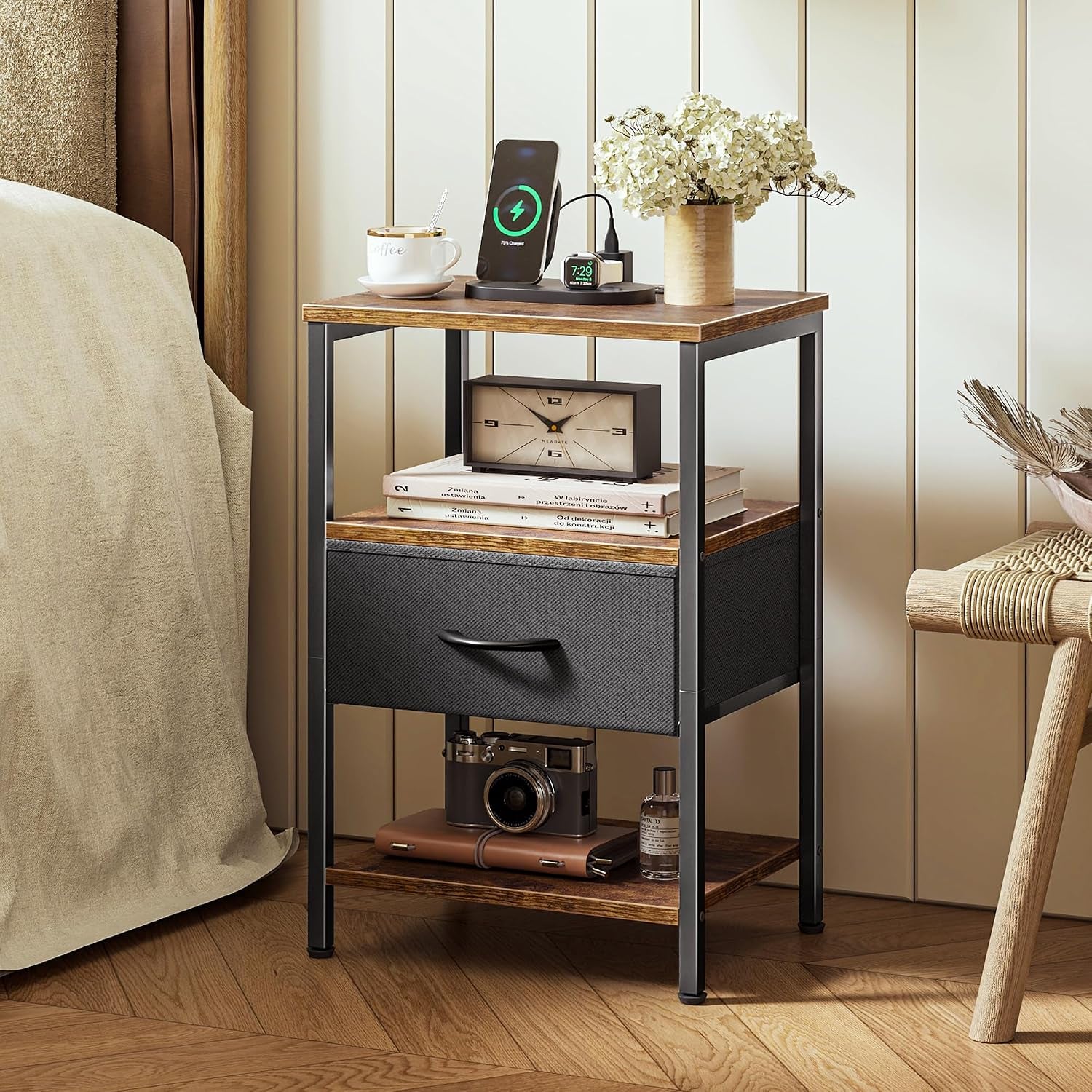 Nightstand styled beside a bed with vase, phone charger and camera on lower shelf in a warm bedroom setting.