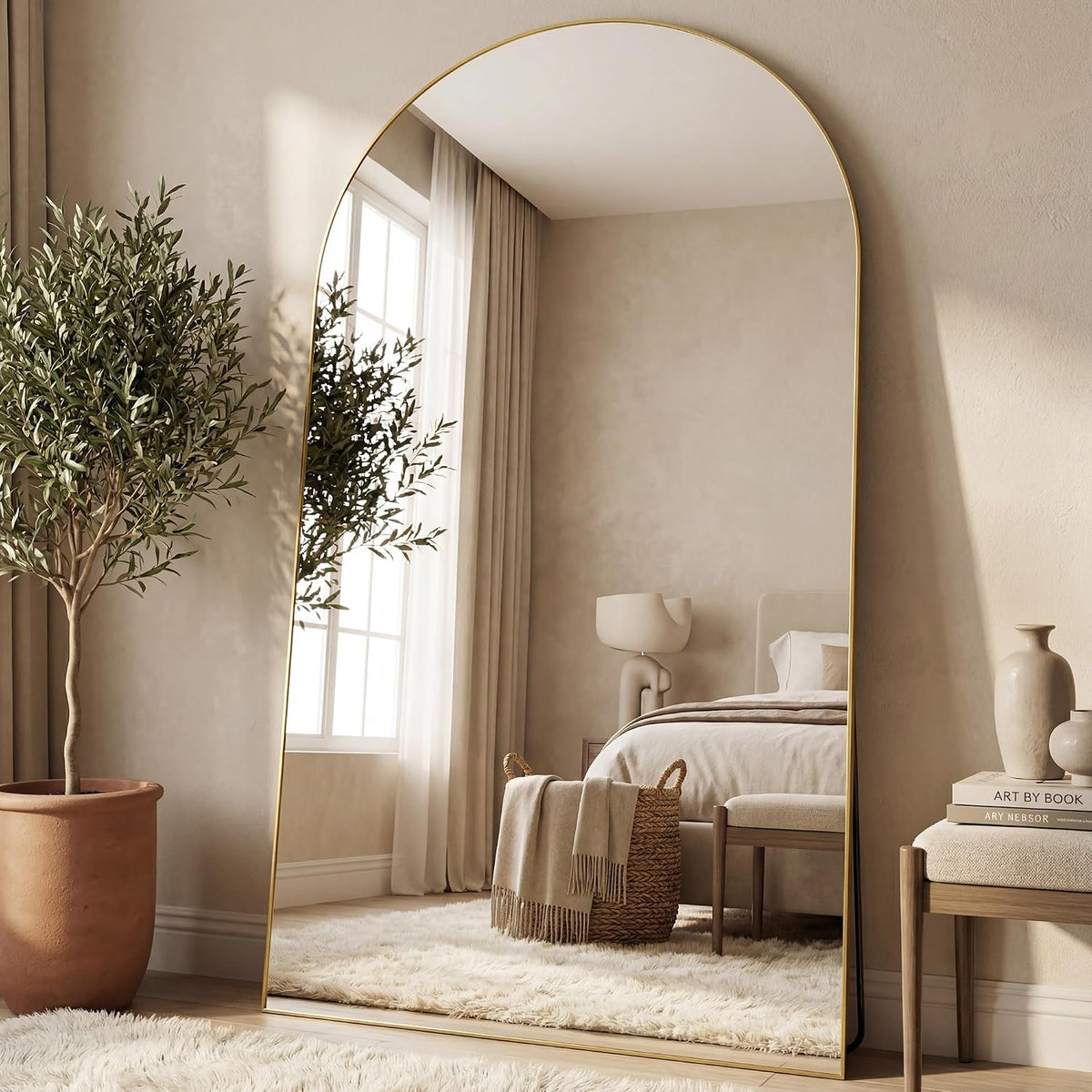 Full-length gold arched mirror propped on floor showing a bed, window, and woven basket in reflection.