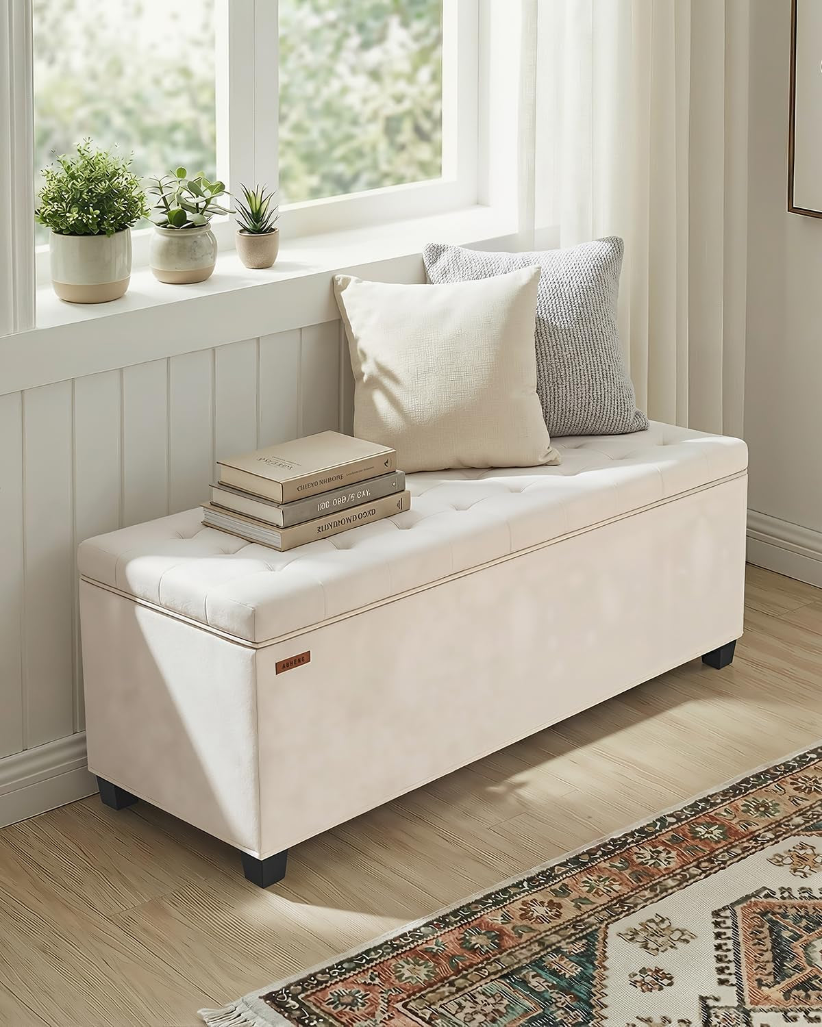 White storage bench by a window topped with pillows and books in a sunlit corner.