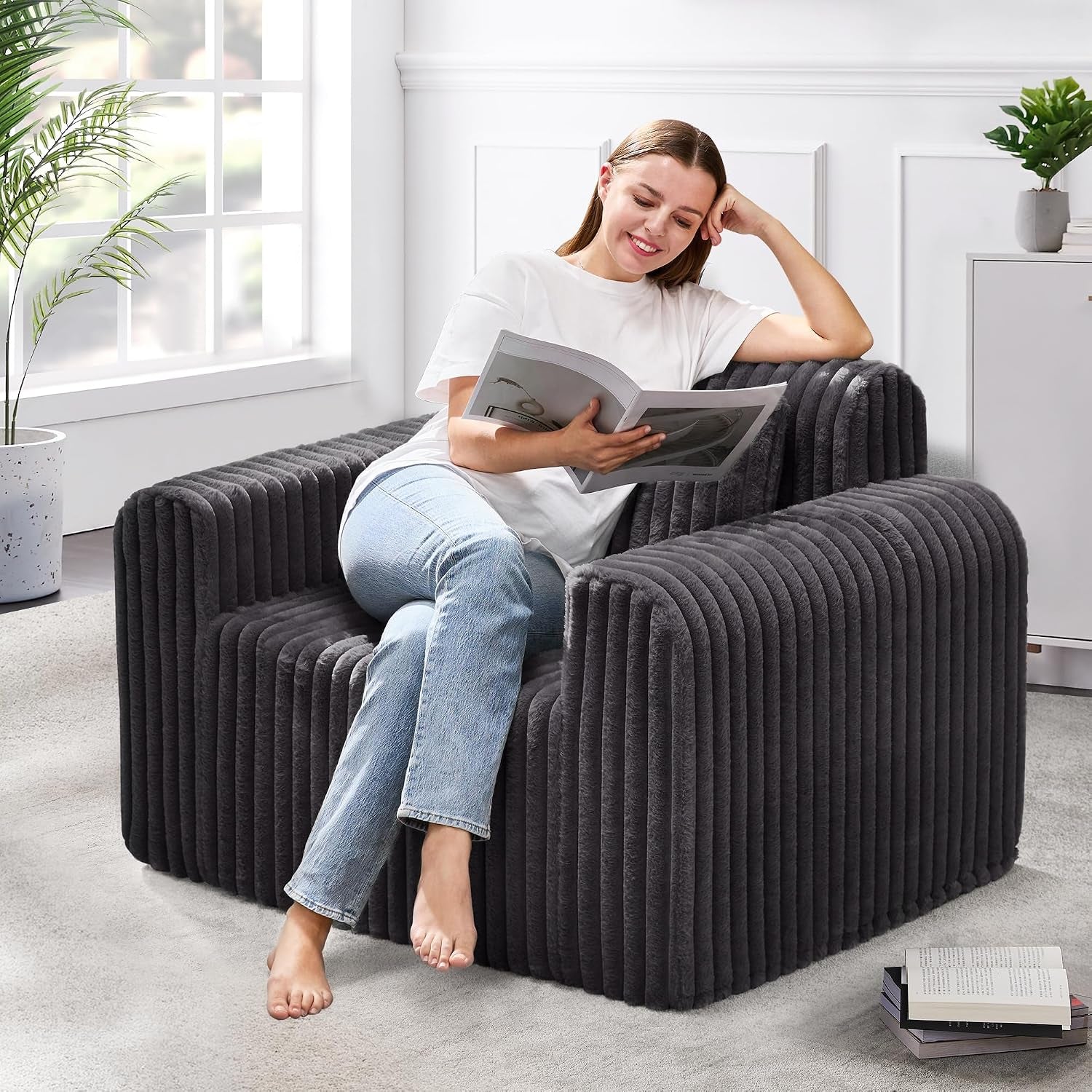 Woman reads on a dark charcoal ribbed floor chair with deep armrests beside a window.