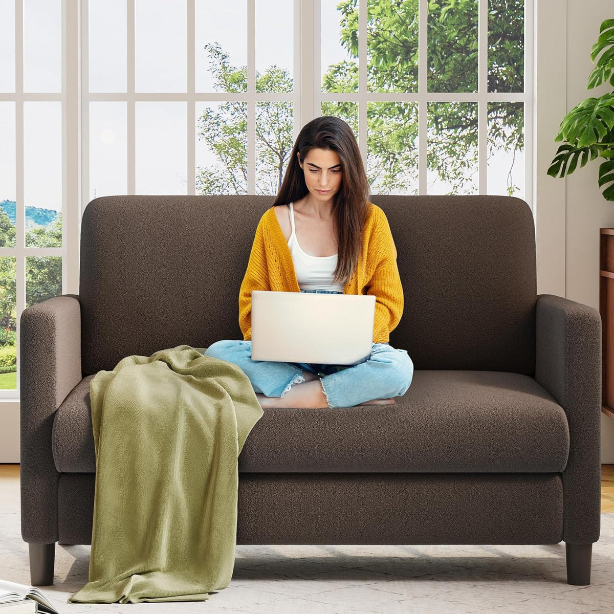 Dark brown two-seat loveseat with woman seated cross-legged using a laptop and a green throw draped over the cushion.