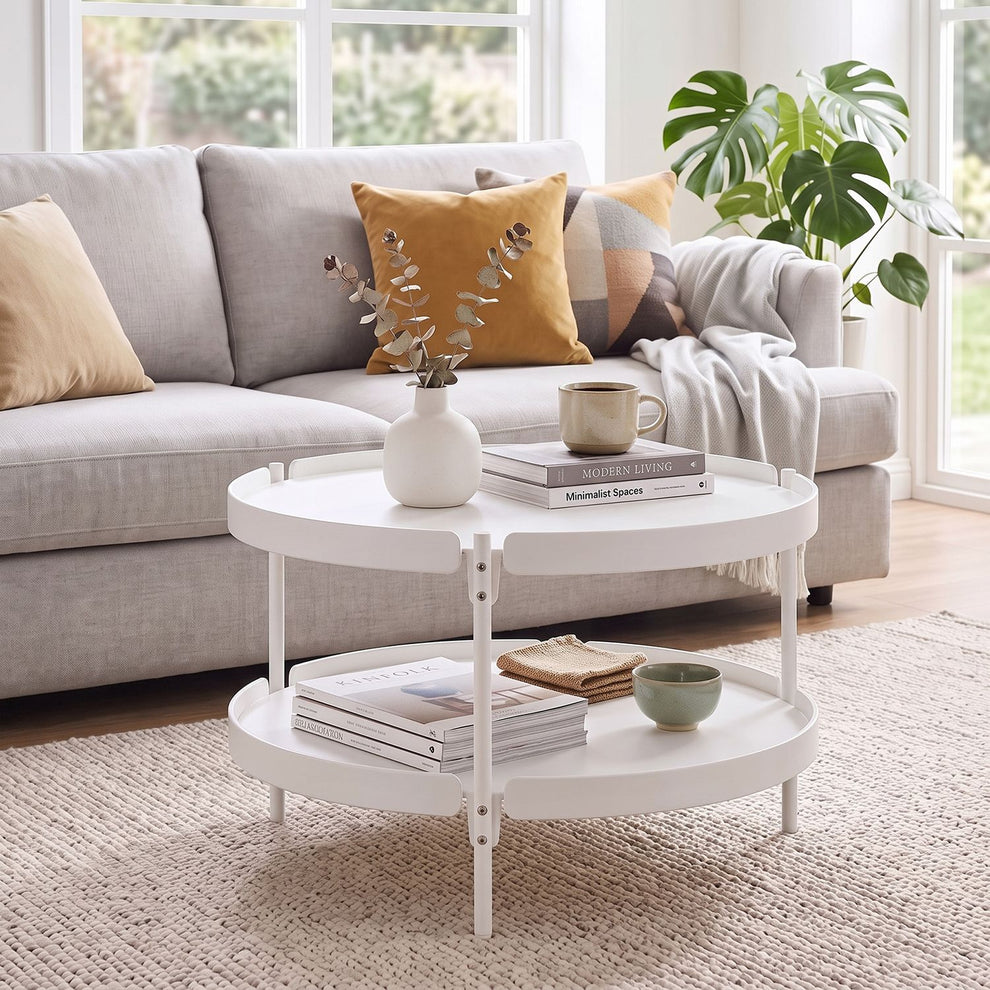 White two-tier round coffee table in front of a light gray sofa, styled with books, a vase, and a cup.