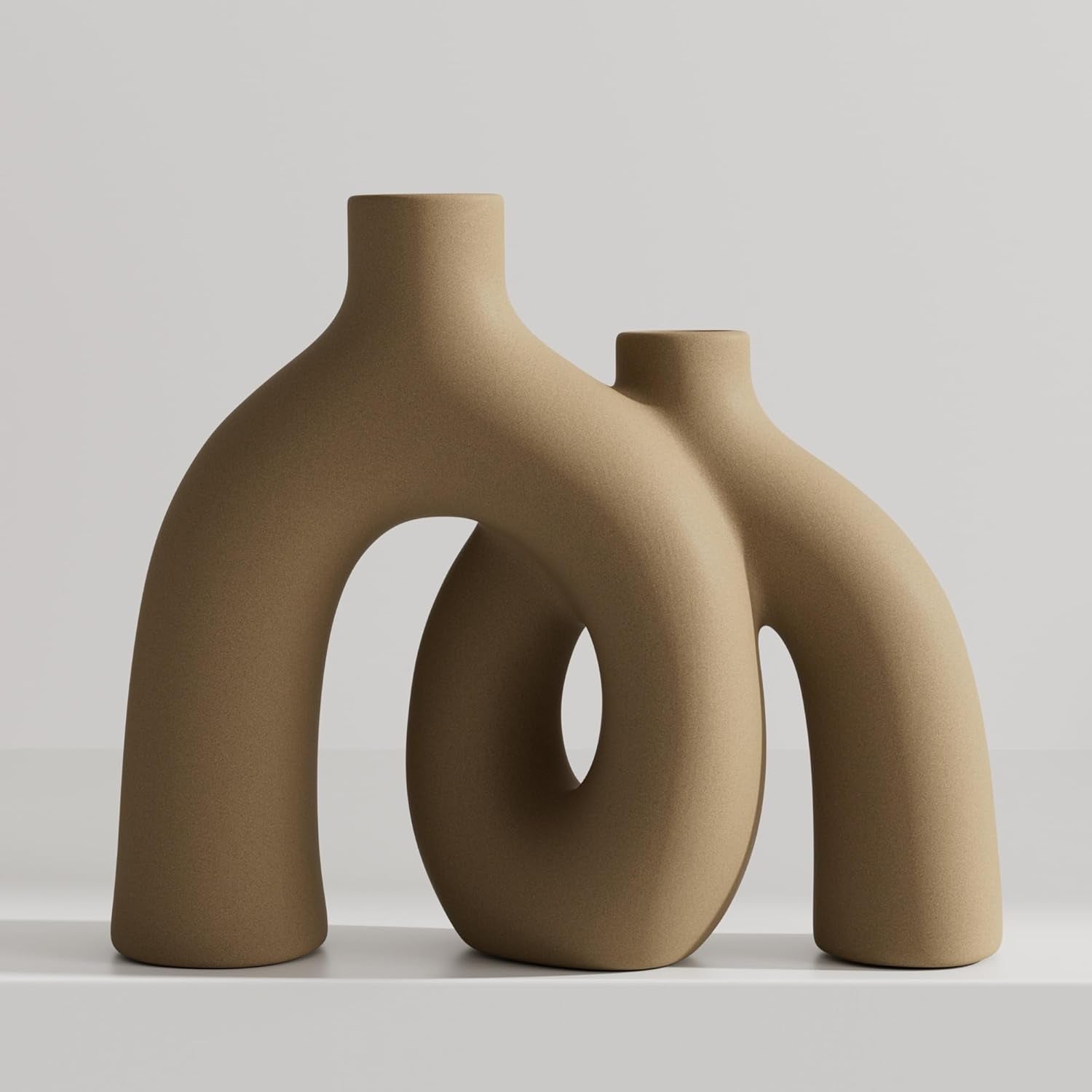 Two interlocking donut-shaped ceramic vases in a matte light tan/beige finish on a white surface.