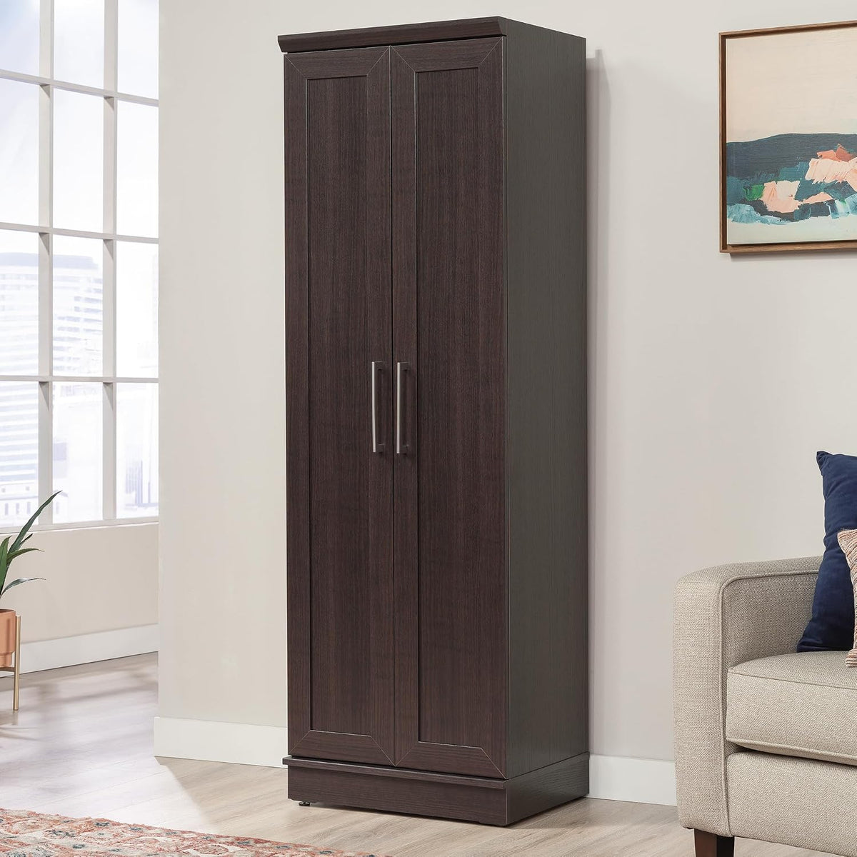Tall two-door dark wood storage cabinet standing against a wall beside a sofa and window in a living room.