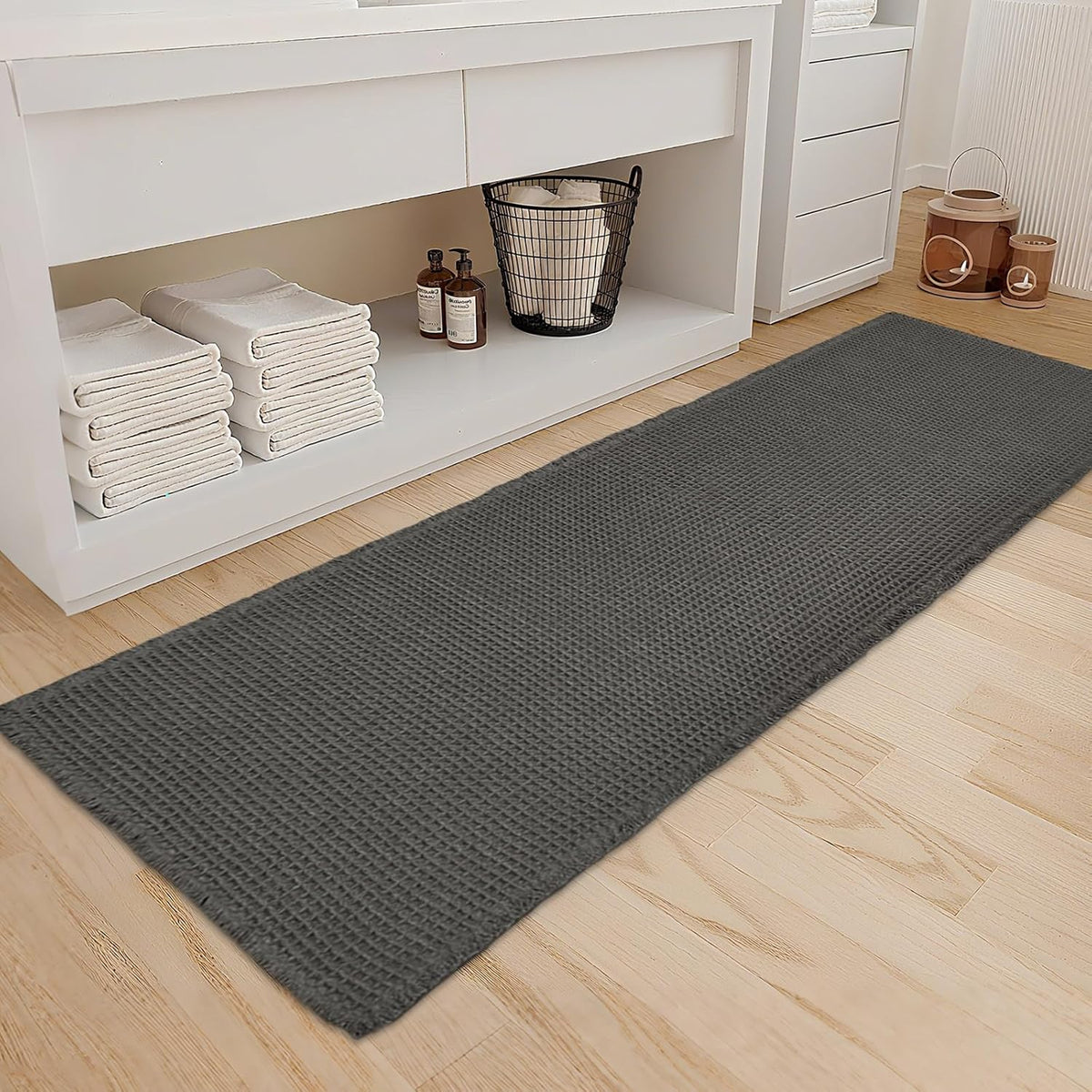 Charcoal-gray waffle runner on a light wood bathroom floor beside open shelving with folded towels and a wire basket.