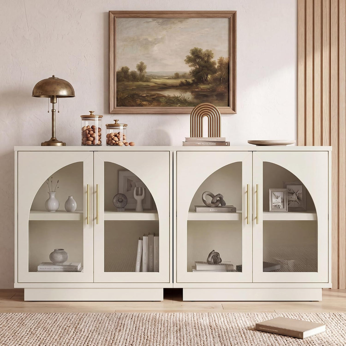 Two white glass-front cabinets side by side with arched door openings, decorated with lamp and artwork.
