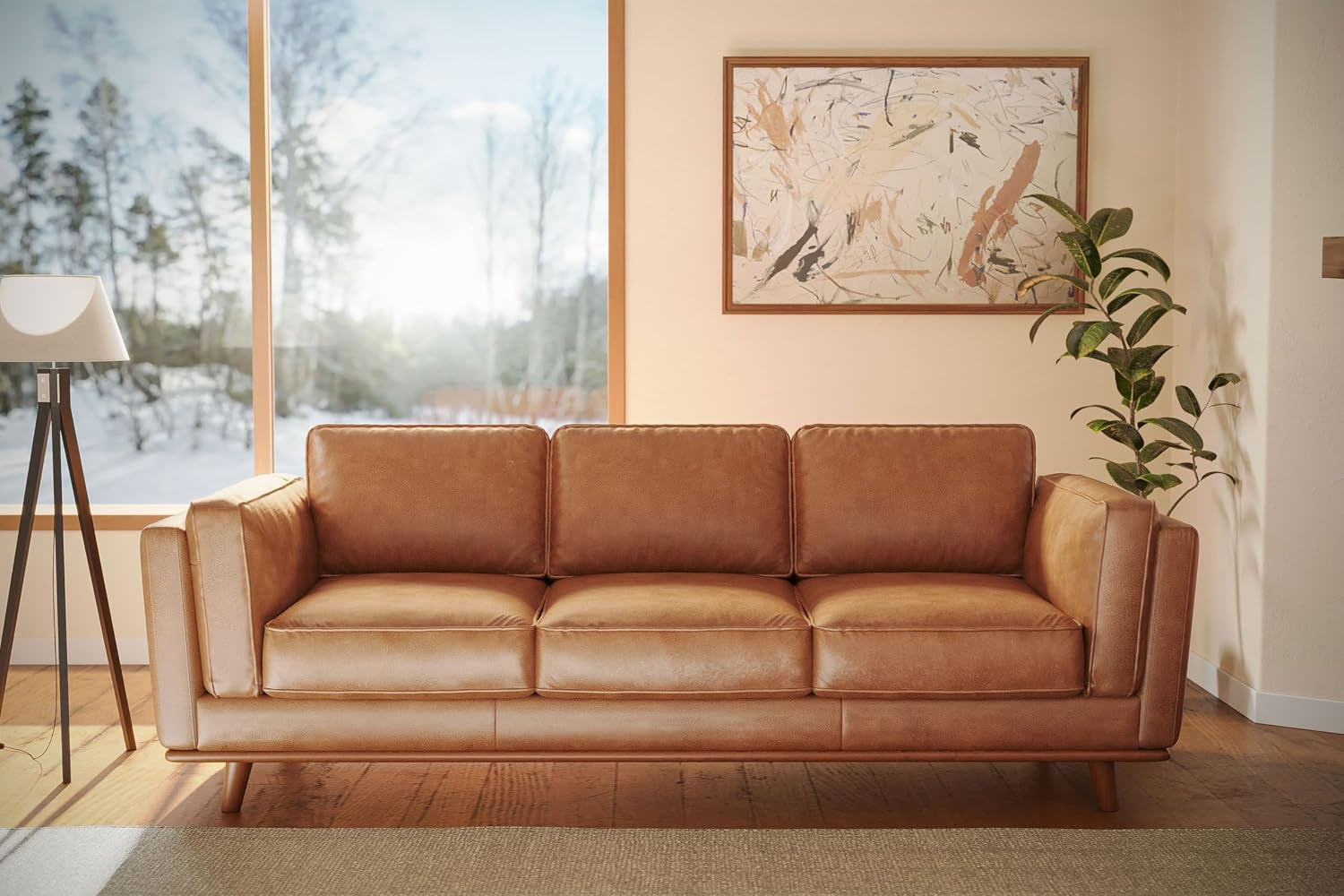 Cognac leather three-seat sofa placed against a wall under a framed painting, flanked by a tripod floor lamp and a tall