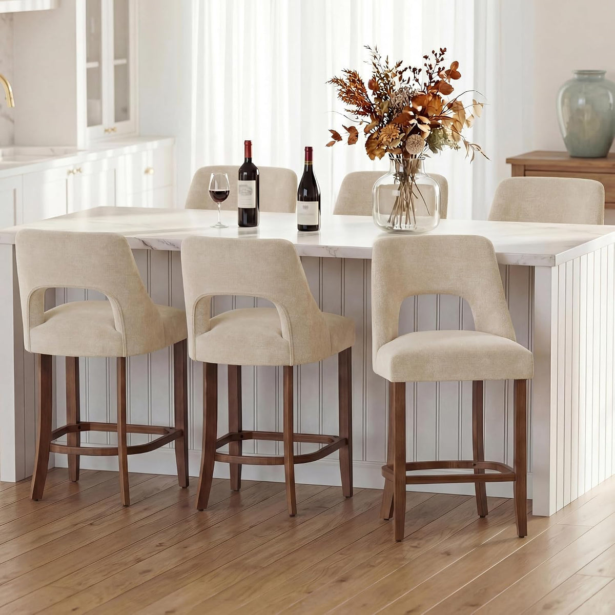 Three beige upholstered counter stools with wood legs tucked at a white island in a bright kitchen