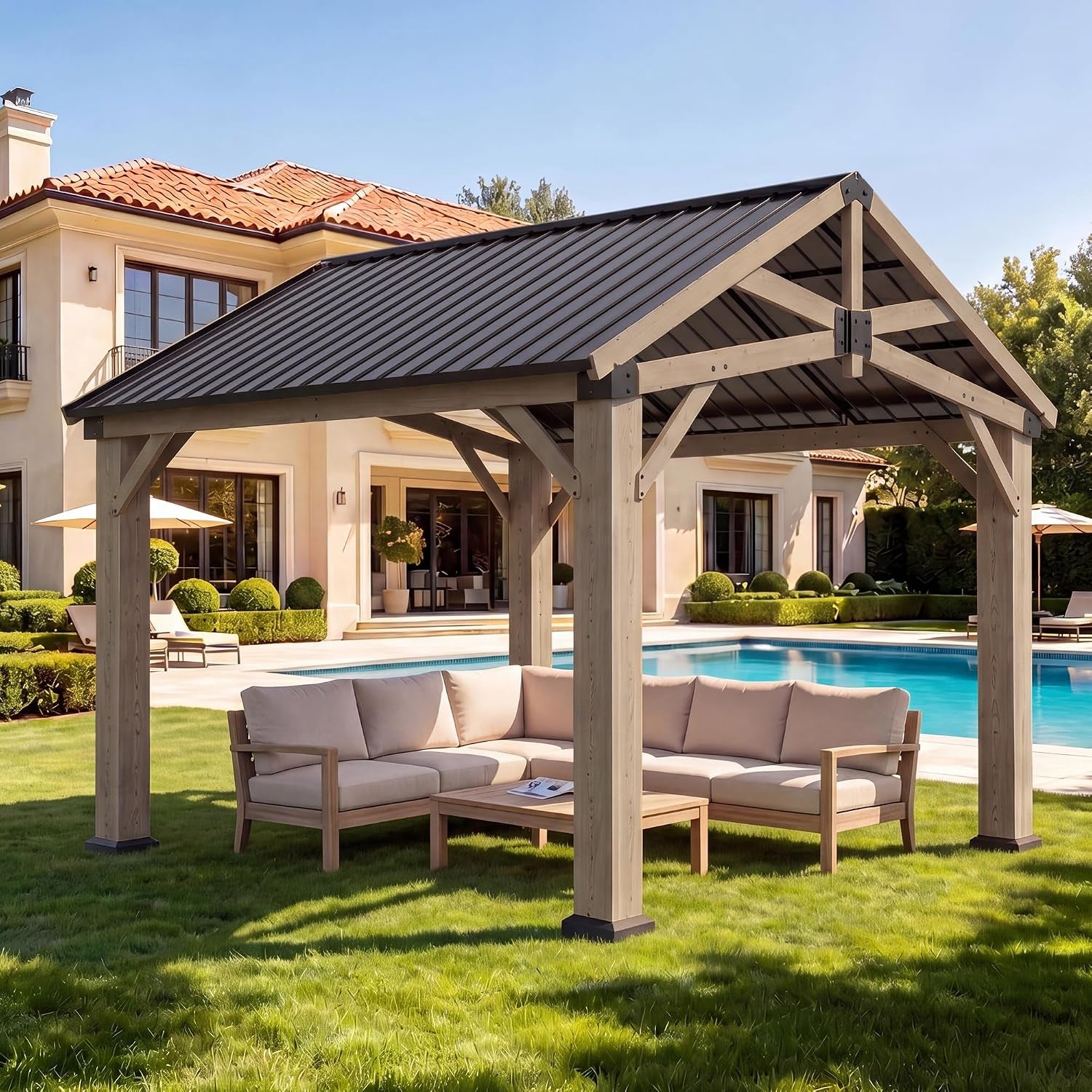 Natural-wood gable gazebo with dark metal roof over a beige sectional by a pool and large stucco house.