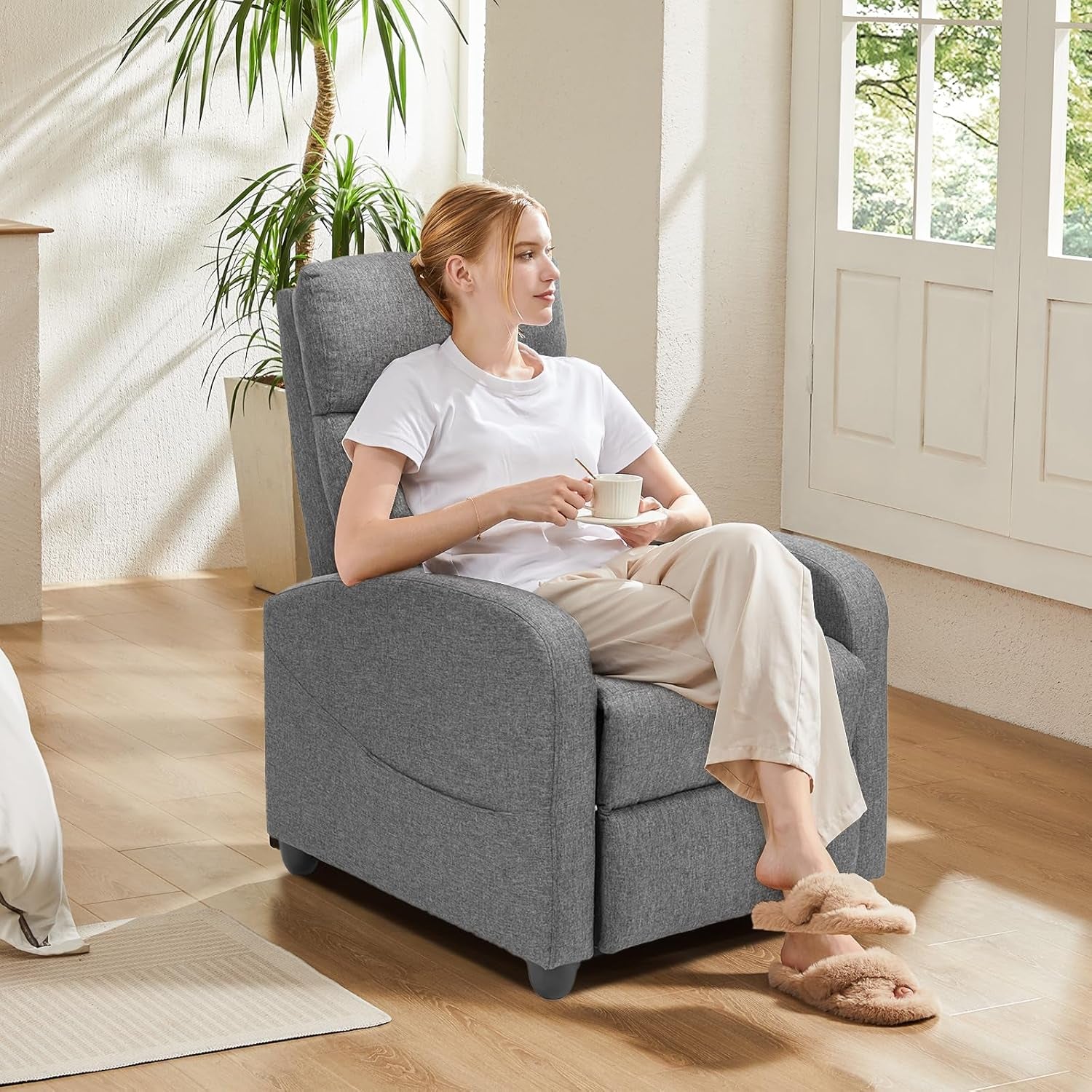 Grey fabric recliner with woman seated upright holding a cup in a bright room by a window
