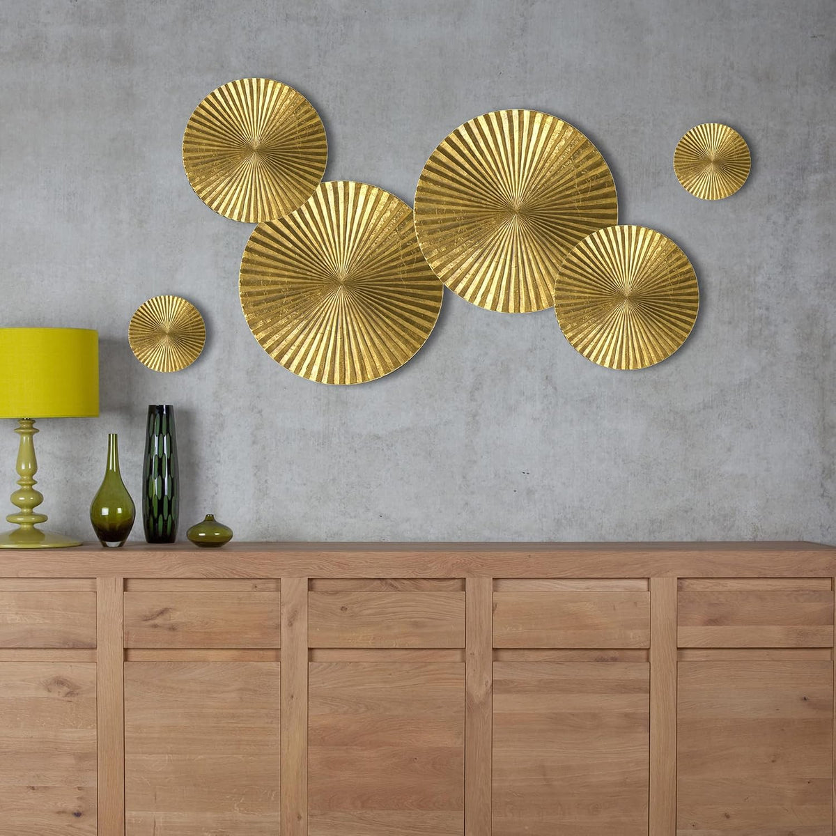 Glossy gold metal sunburst wall art set mounted on a gray wall beside a lamp and decorative vases on a wood console