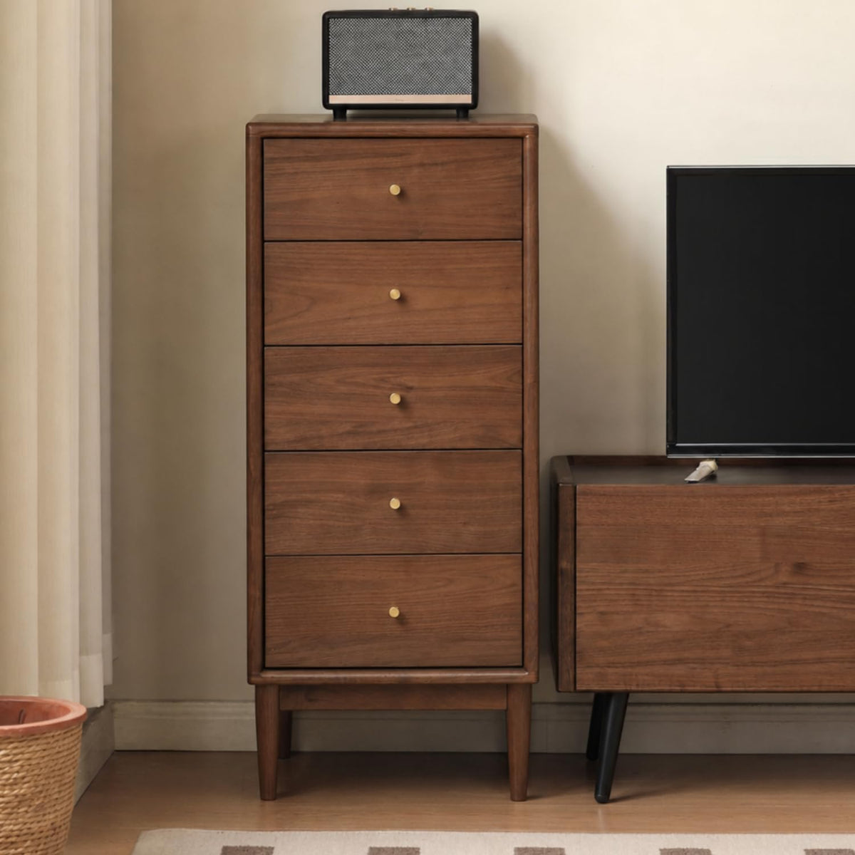 Side view of the tall narrow chest in a darker wood next to a low media cabinet and a small speaker on top.