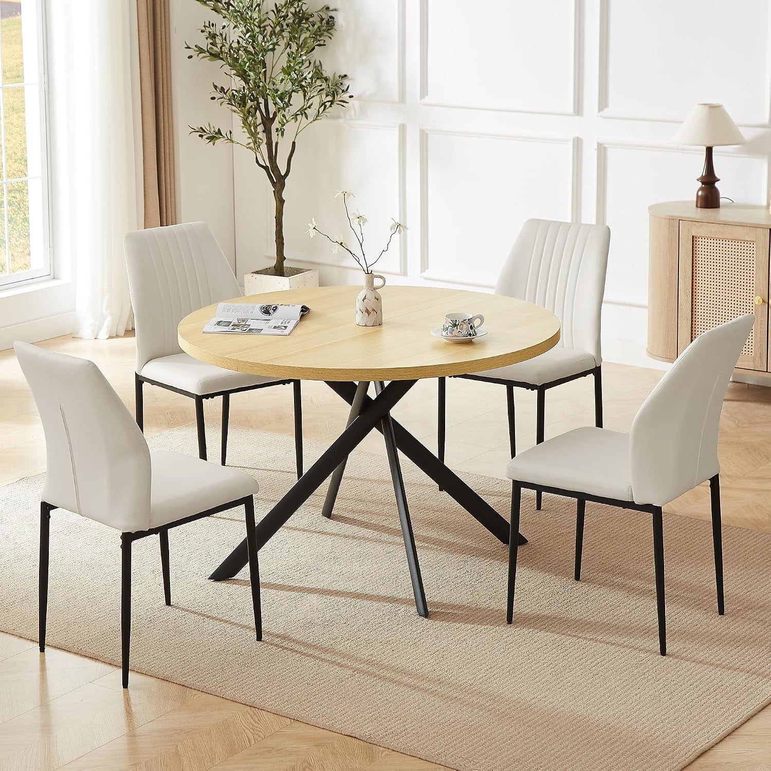 Round light-wood dining table on black crisscross legs with four smooth white chairs having thin black legs.
