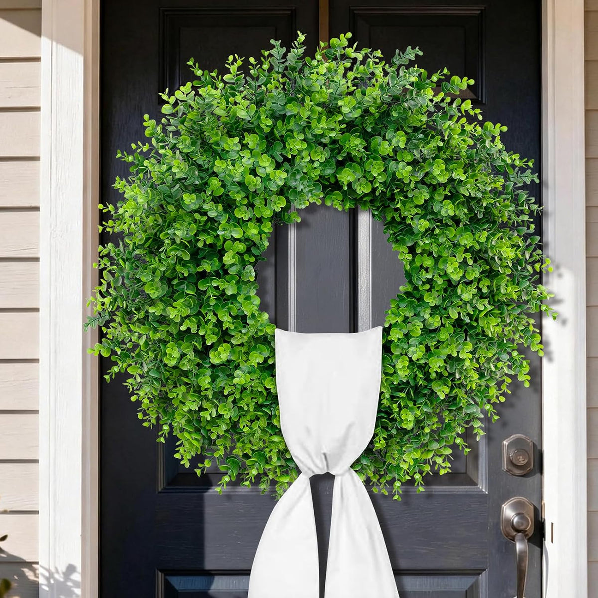 Close-up of a dense green boxwood wreath showing small rounded leaves and a white sash over a dark door.