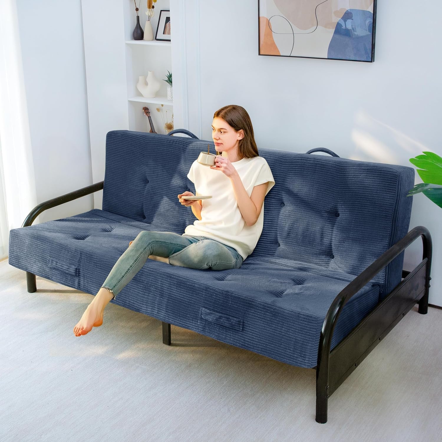 Woman sitting on a dark blue corded-fabric futon mattress on a black metal frame in a bright living room.