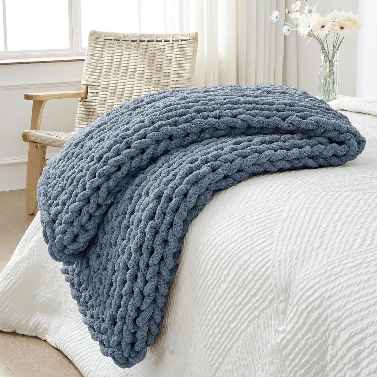 Blue-gray chunky knit throw folded and draped over a white quilted bed with a woven chair and flowers nearby.