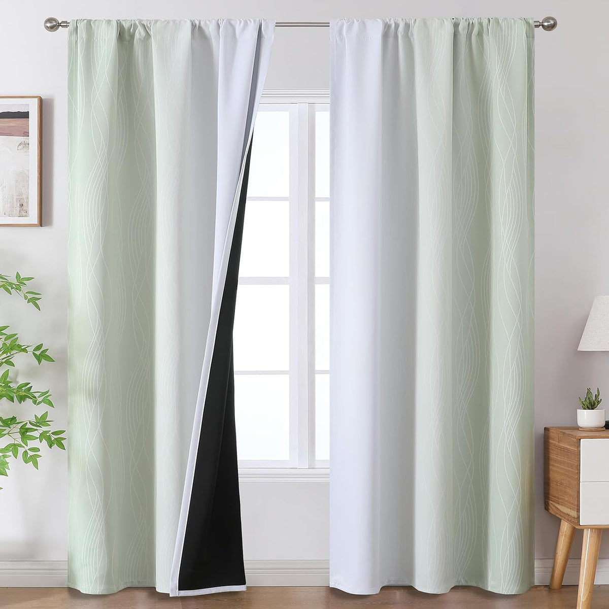Pair of light green-to-white ombre curtains with black blackout backing hung on a window, bedside table and plant visible.