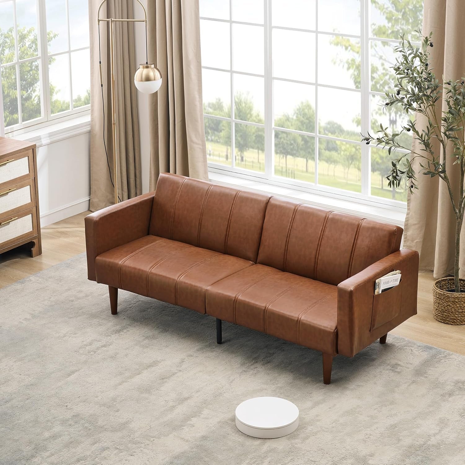 Angled room view of brown PU leather futon sofa placed by a large window with a neutral rug beneath