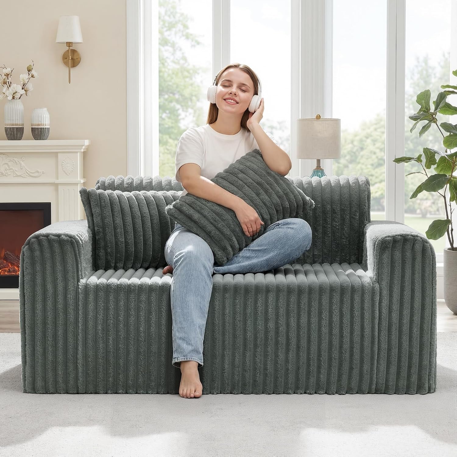 Woman with headphones sits on a ribbed moss-gray floor sofa holding a matching cushion in a bright room.