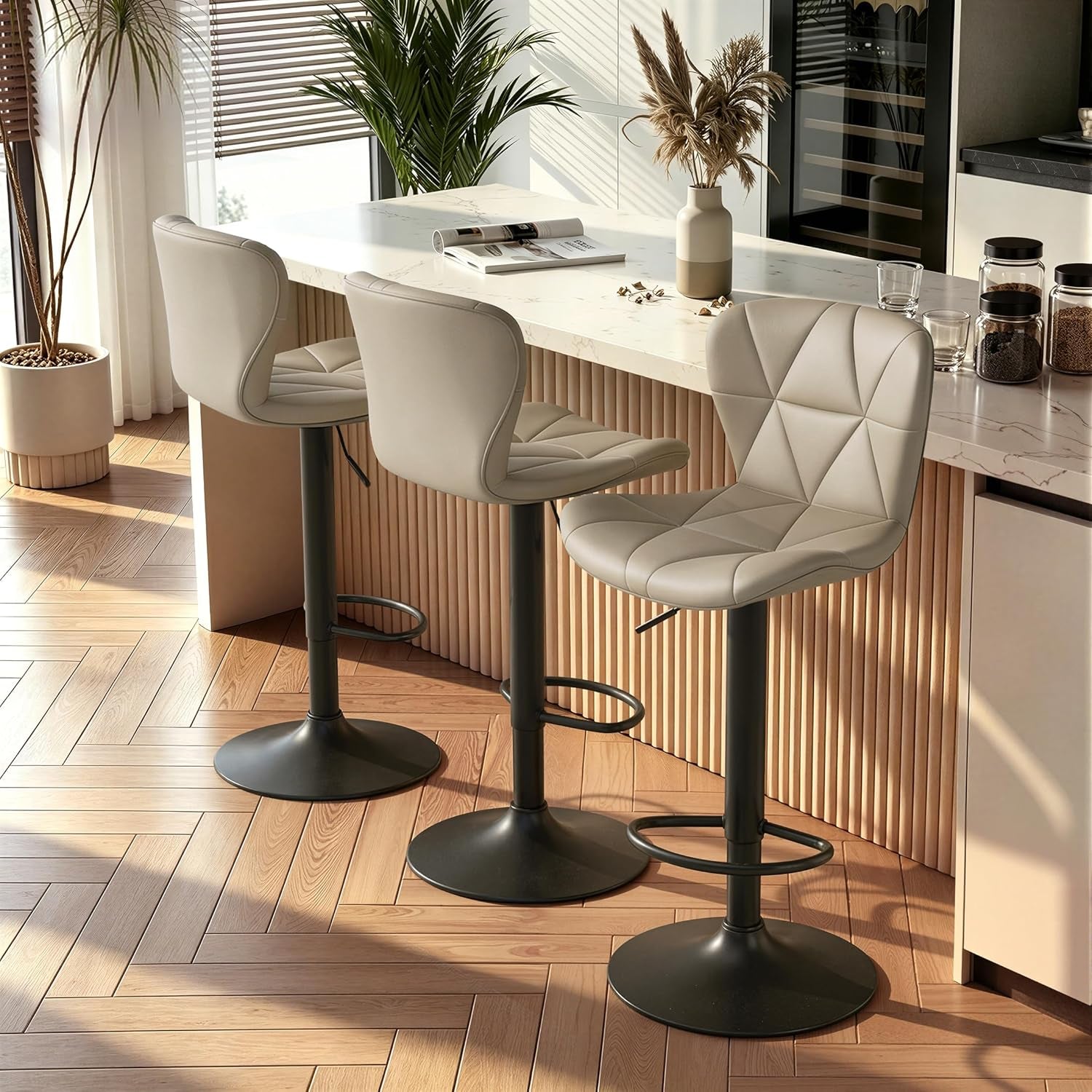 Three light-gray bar stools in a row at a fluted-front counter with sunlight and houseplants nearby.