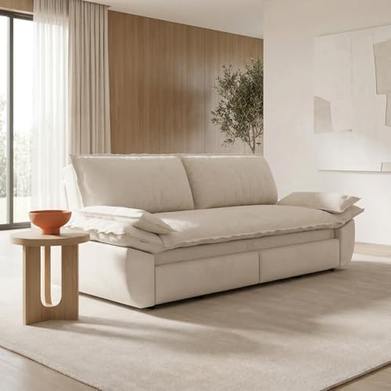 Side view of the beige sofa in seating mode with plush back cushions, a folded throw, and a small round wooden side table.