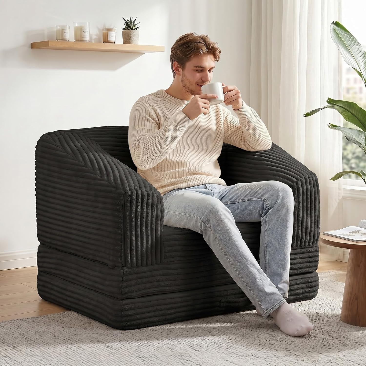Man sipping coffee while seated in a black corduroy single-seat folding sofa chair in a bright room.
