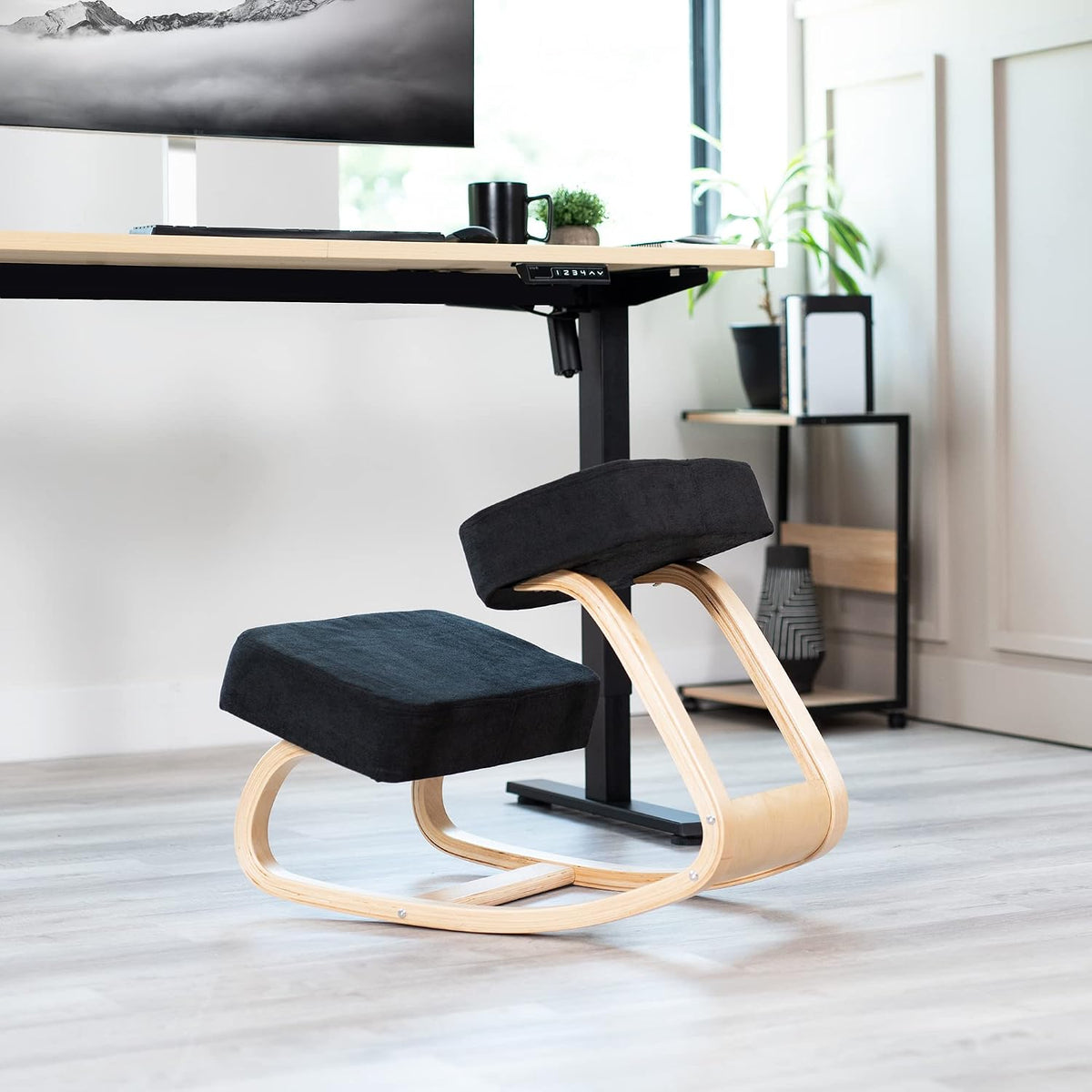 Black-cushioned kneeling chair with light wood rocker base beside a standing desk in a bright home office