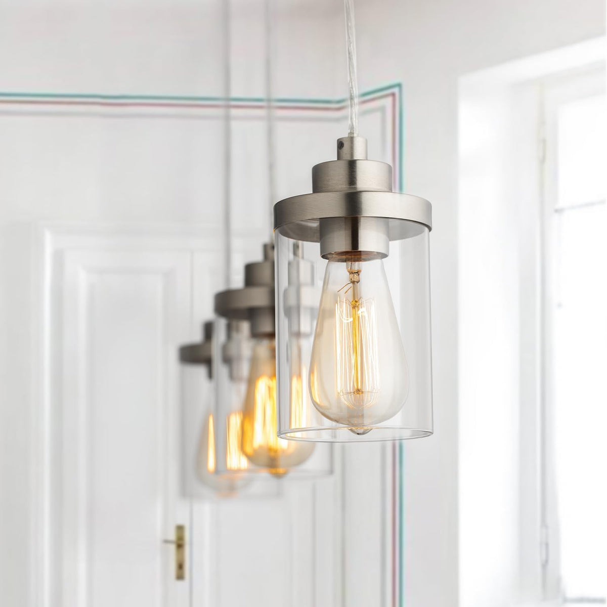 Close-up of a brushed-nickel pendant with clear glass cylinder and a glowing filament bulb, angled front view