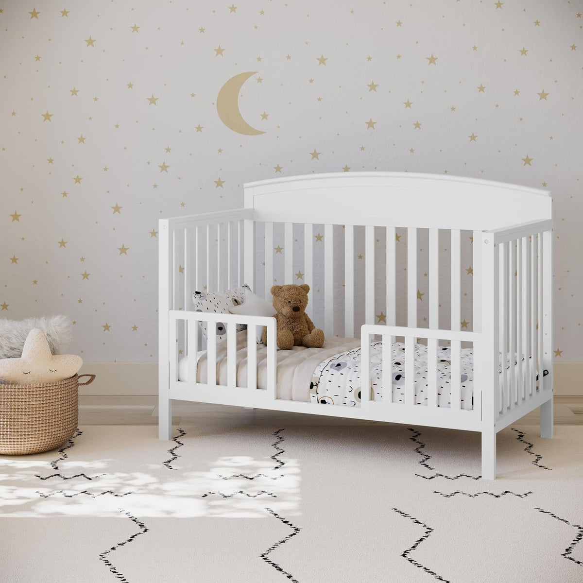 Crib configured as a toddler bed with low front rail, stuffed bear and pillows inside, star-patterned wall behind