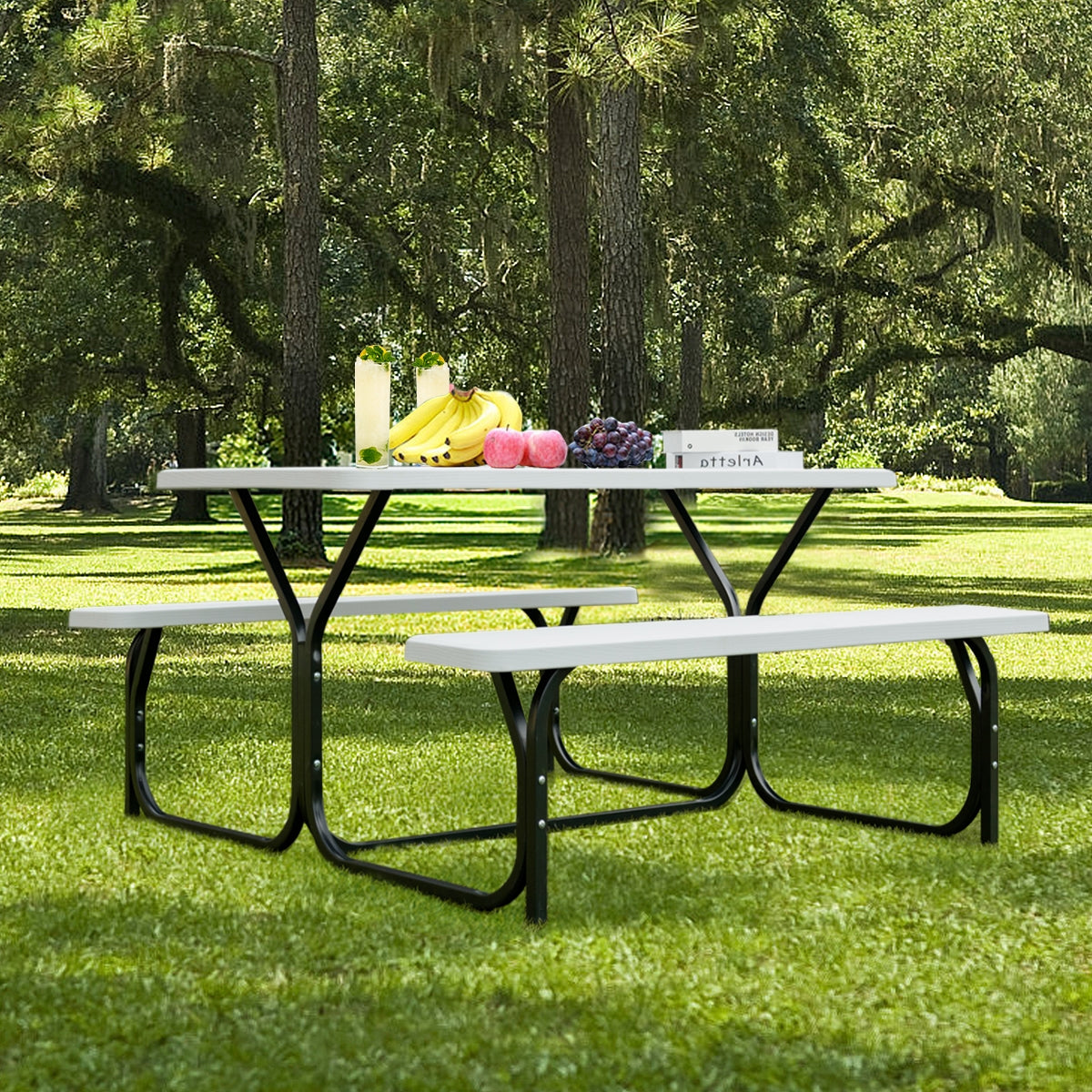 Gray picnic table and two benches on a lawn beneath leafy trees, with fruit and drinks on the tabletop.