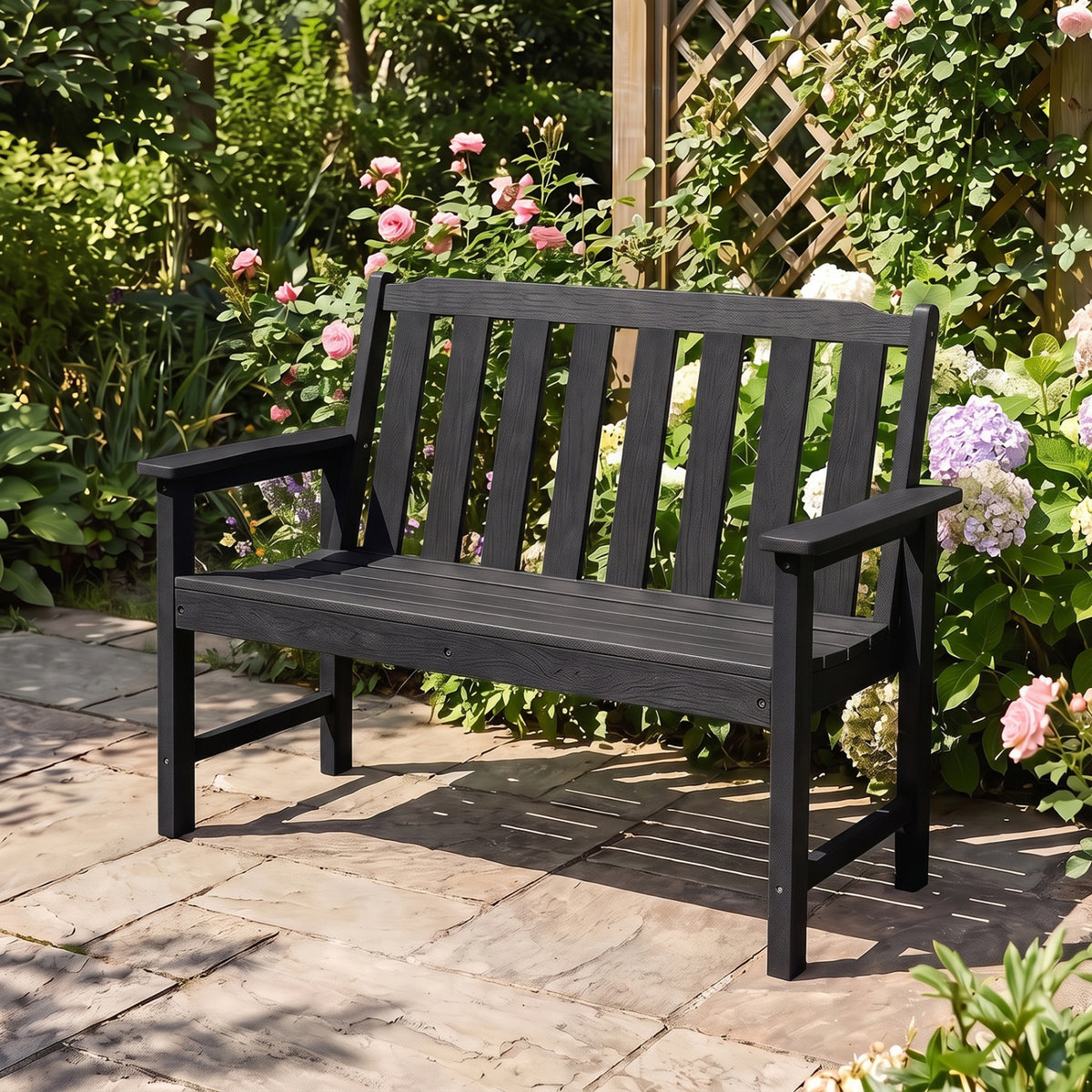Black slatted garden bench with a tall back and armrests, surrounded by flowering plants.