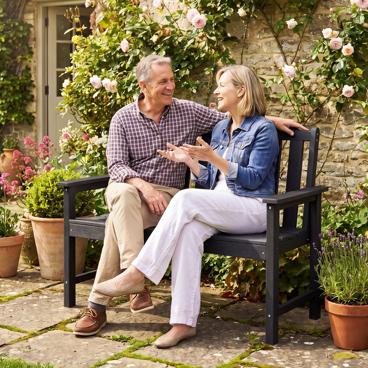 A smiling couple sits together on a black slatted bench in a flower-filled garden.