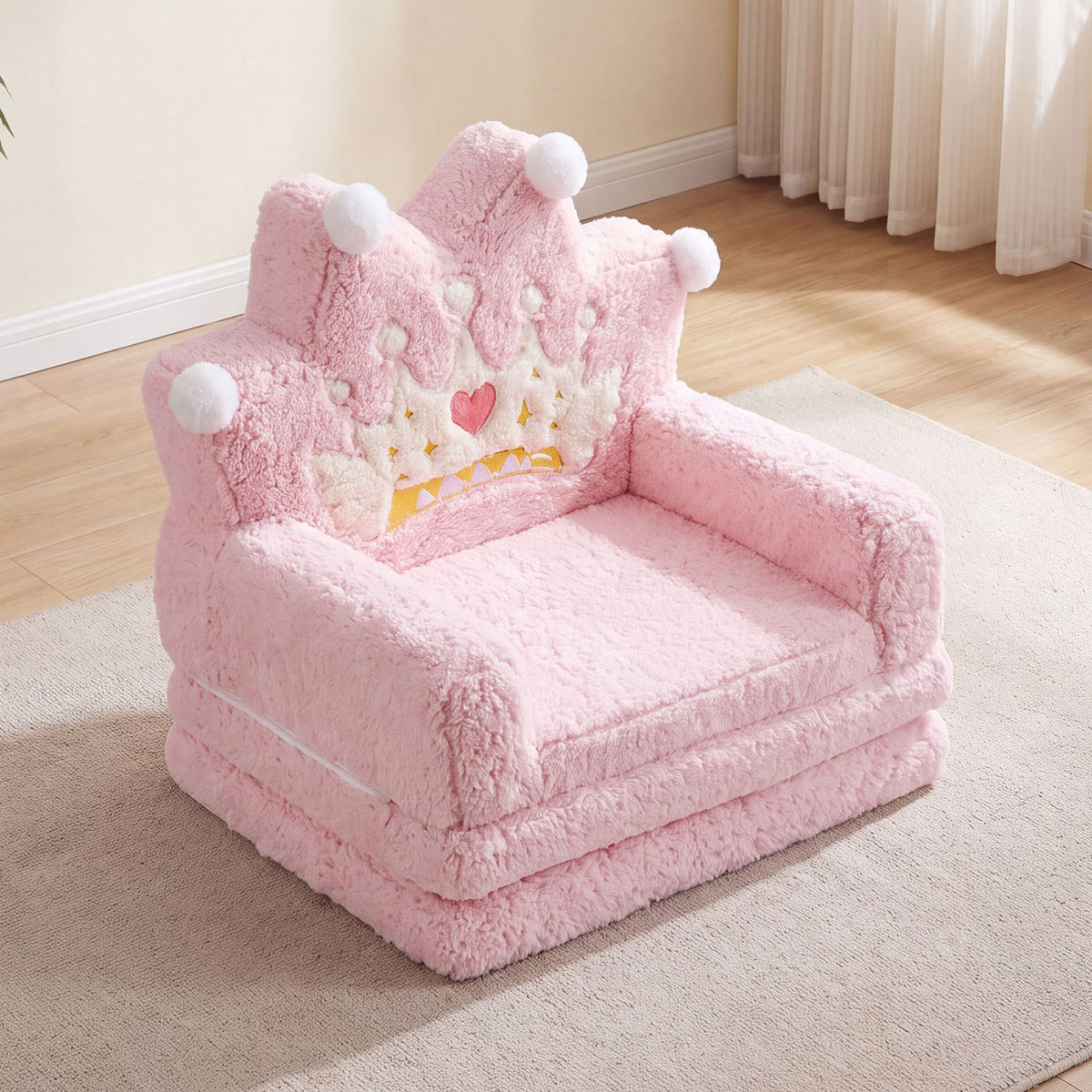 Pink plush children’s sofa with a crown-shaped back, white pom-poms and a heart motif, set on a rug.