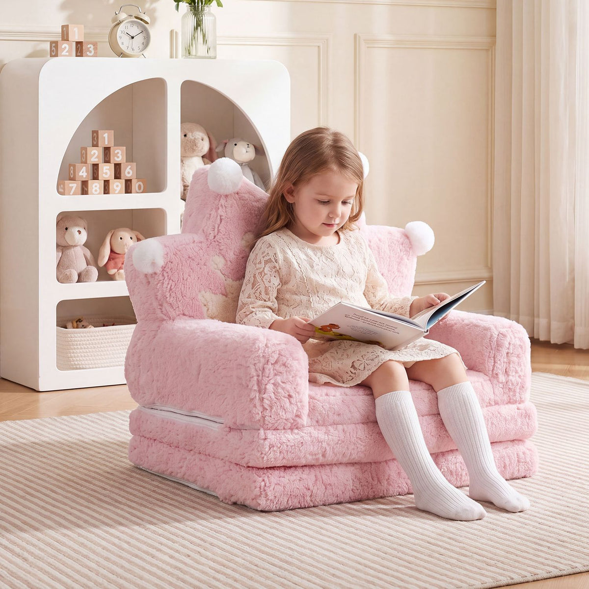 A young girl reads while seated on a pink plush kids sofa in a bright room.