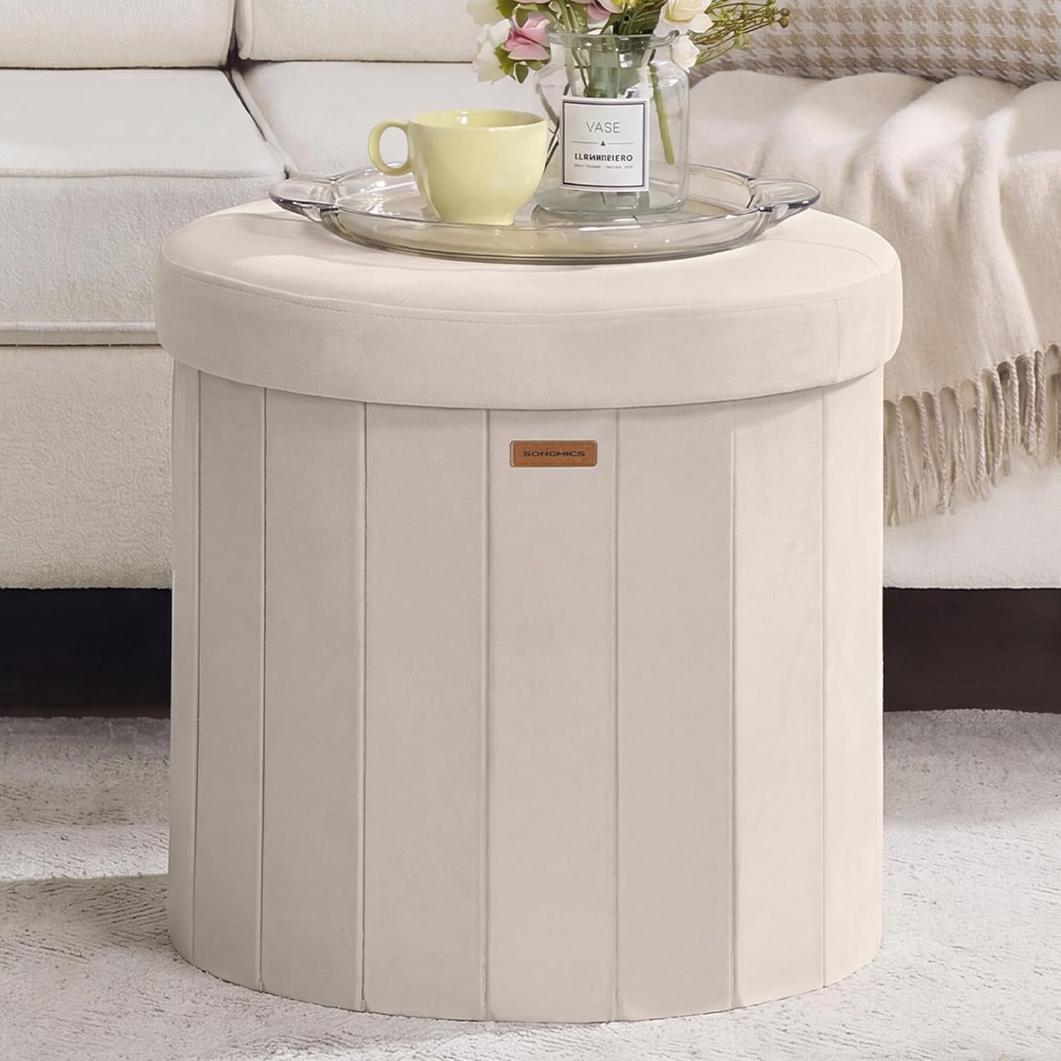 Beige round velvet storage ottoman shown front view beside a sofa with a tray on top