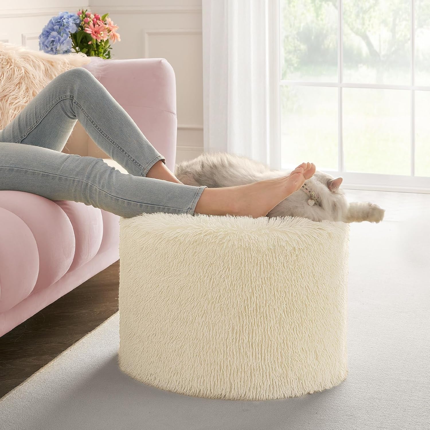 Side living room shot of a cream round pouf used as a footrest with a person’s legs on it and a fluffy cat resting nearby.