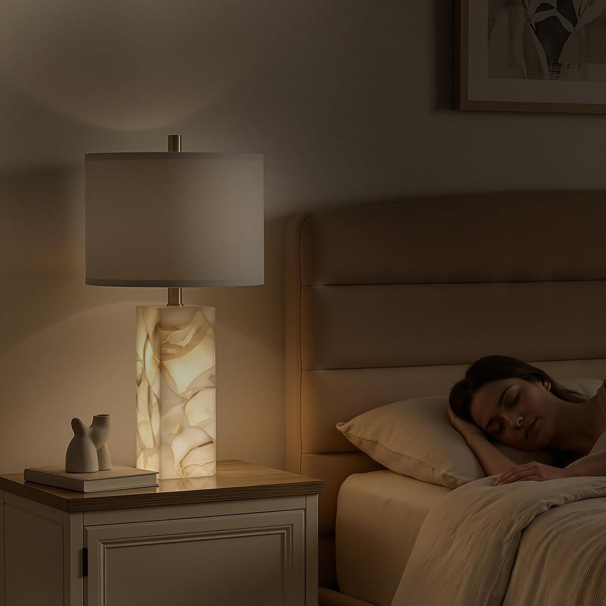 Lit alabaster column bedside lamp with white linen shade on a nightstand beside a sleeping person