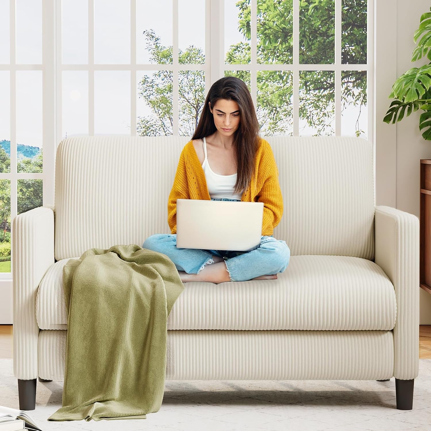 Woman on a cream ribbed-texture loveseat working on a laptop with a draped green throw
