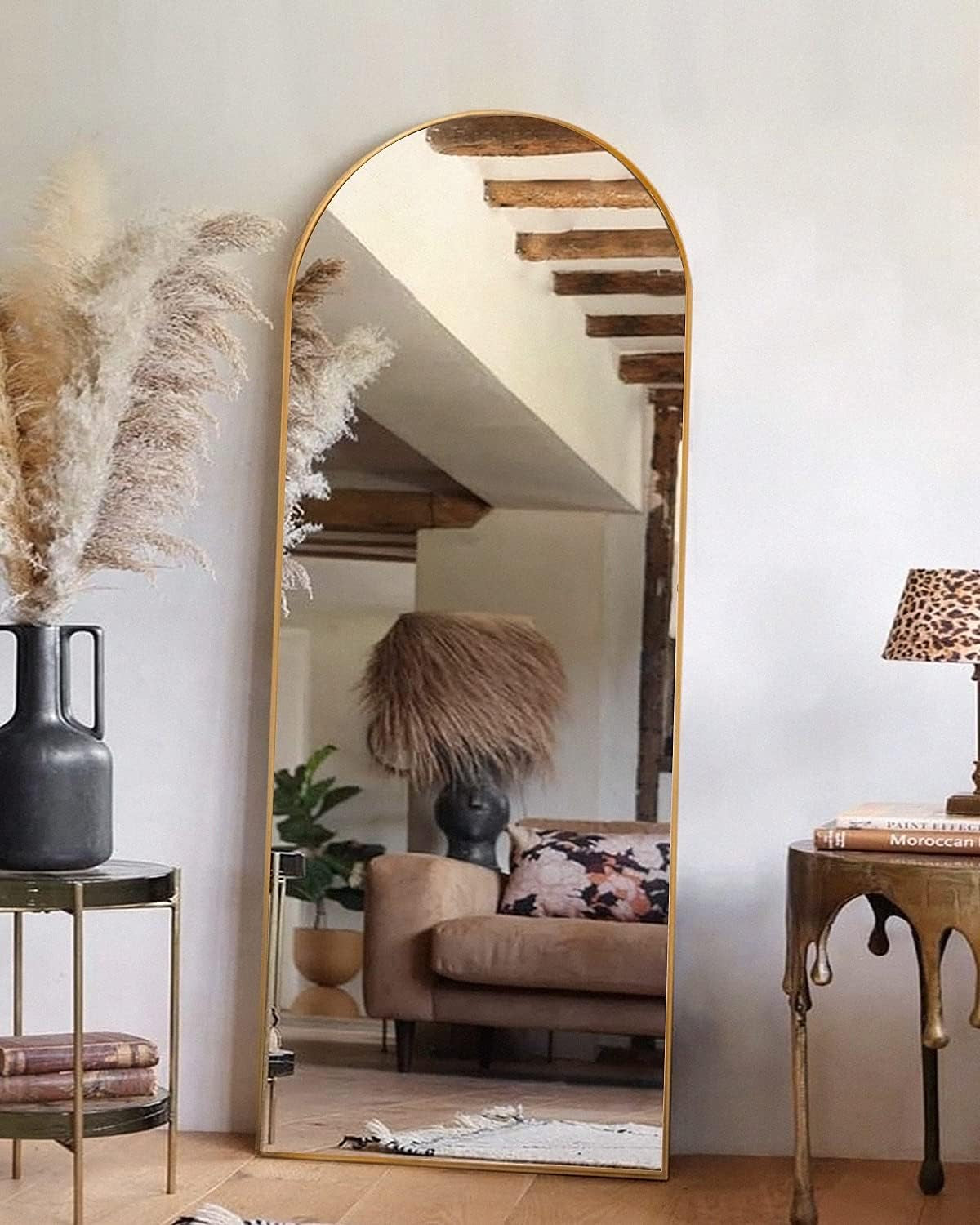 Arched full-length mirror with thin gold frame leaning against a wall, reflecting a sofa and decorative lamp.