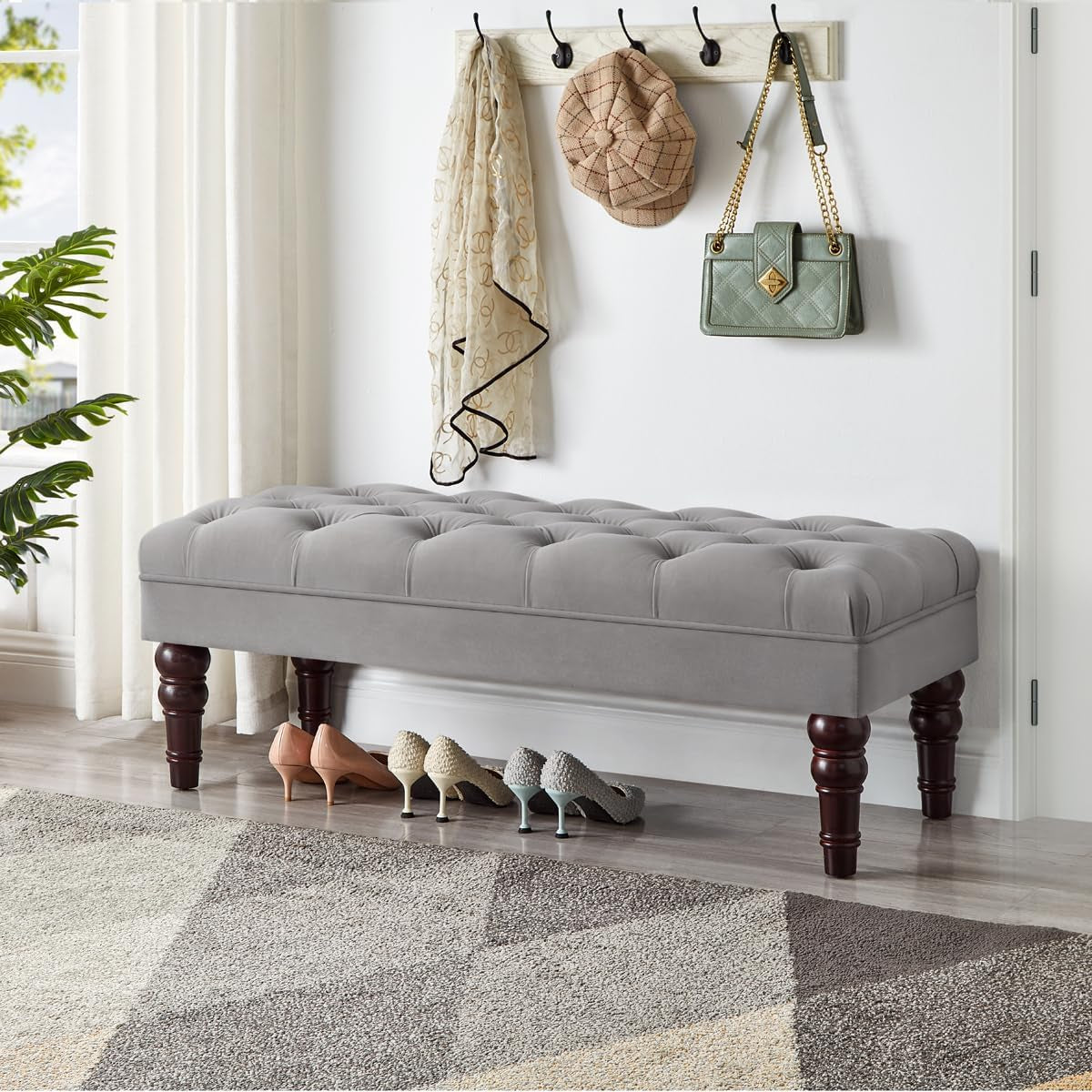 Light gray tufted bench with turned dark-wood legs beneath wall hooks, purse and scarves above