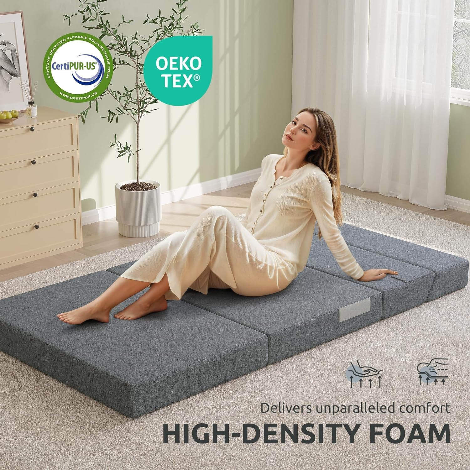 Person seated on unfolded dark gray mattress laid flat on carpet demonstrating lounge use
