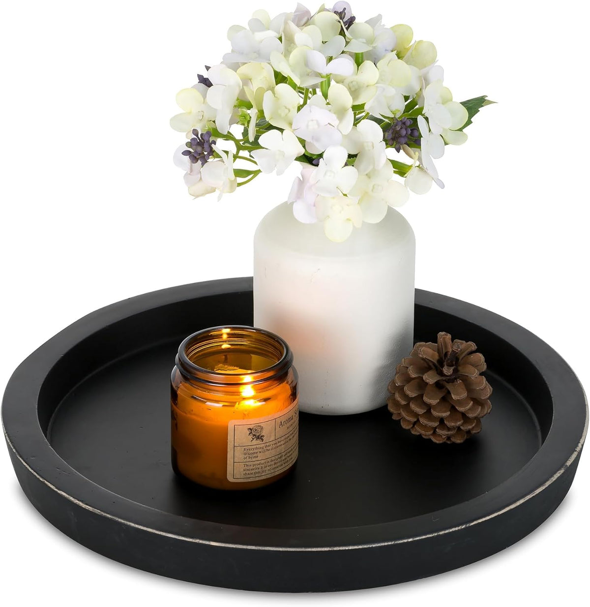 Black round tray holding a lit amber jar candle, white vase, and a pinecone on a white background.