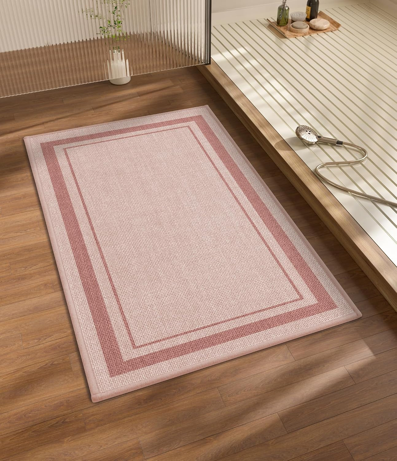Rectangular pink bath rug with double border positioned on wood floor near a tub and shower head.