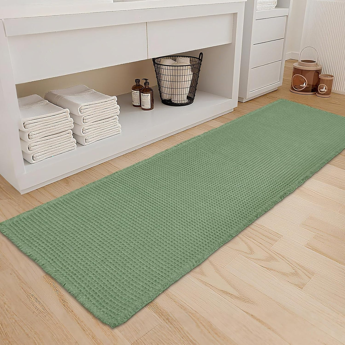 Long waffle-textured bath mat in a soft sage green laid on light wood floor beside white cabinet.