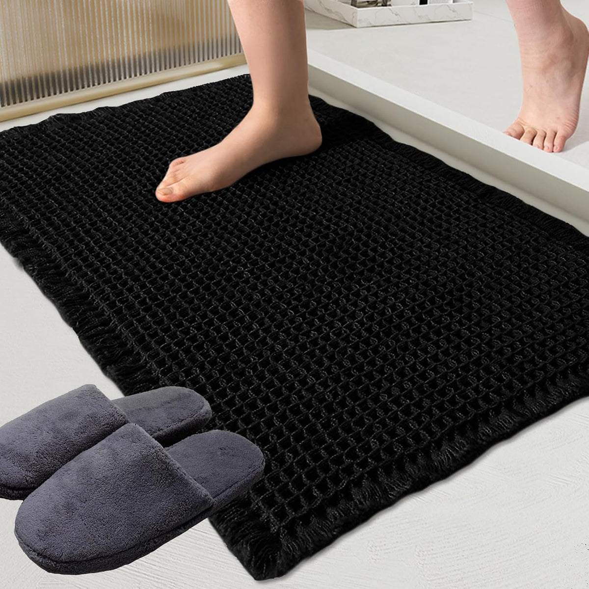 Close-up of a black waffle-texture bath mat with a bare foot stepping onto it and gray slippers nearby