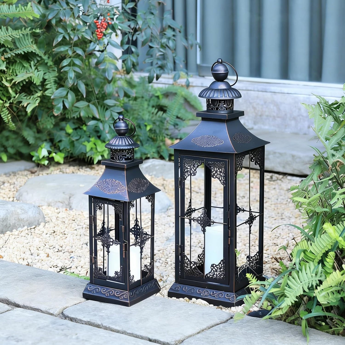 Two ornate black metal lanterns with pillar candles sitting on gravel among ferns and stepping stones.