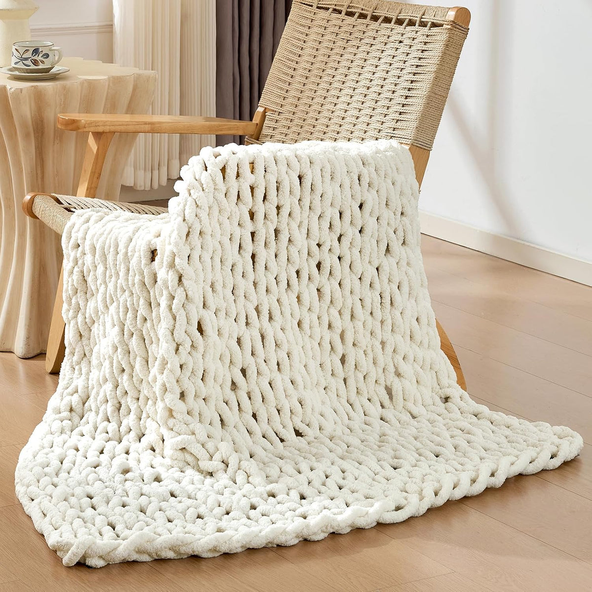 Cream chunky knit throw casually draped over a woven chair beside a sculpted side table.